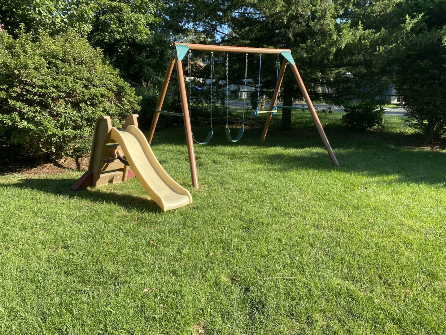 A wooden swing set with a slide stands on a green lawn under trees.